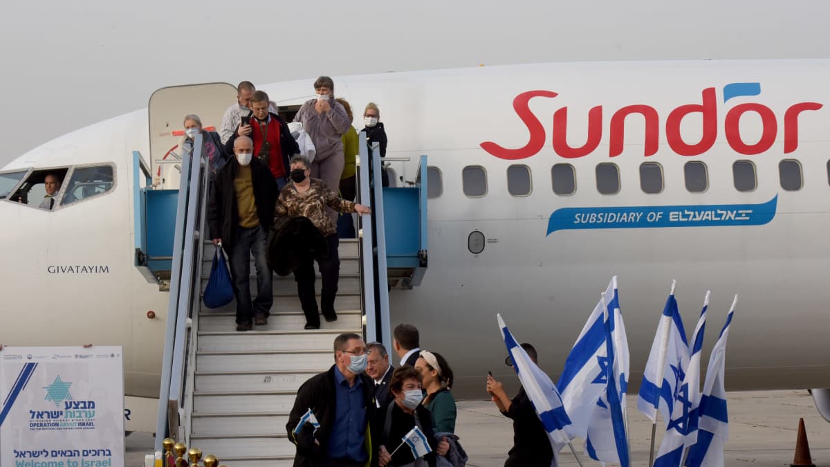Ihmisiä nousee lentokoneesta portaita alas. Maassa on punainen matto ja telineessä Israelin valkosinisiä lippuja.