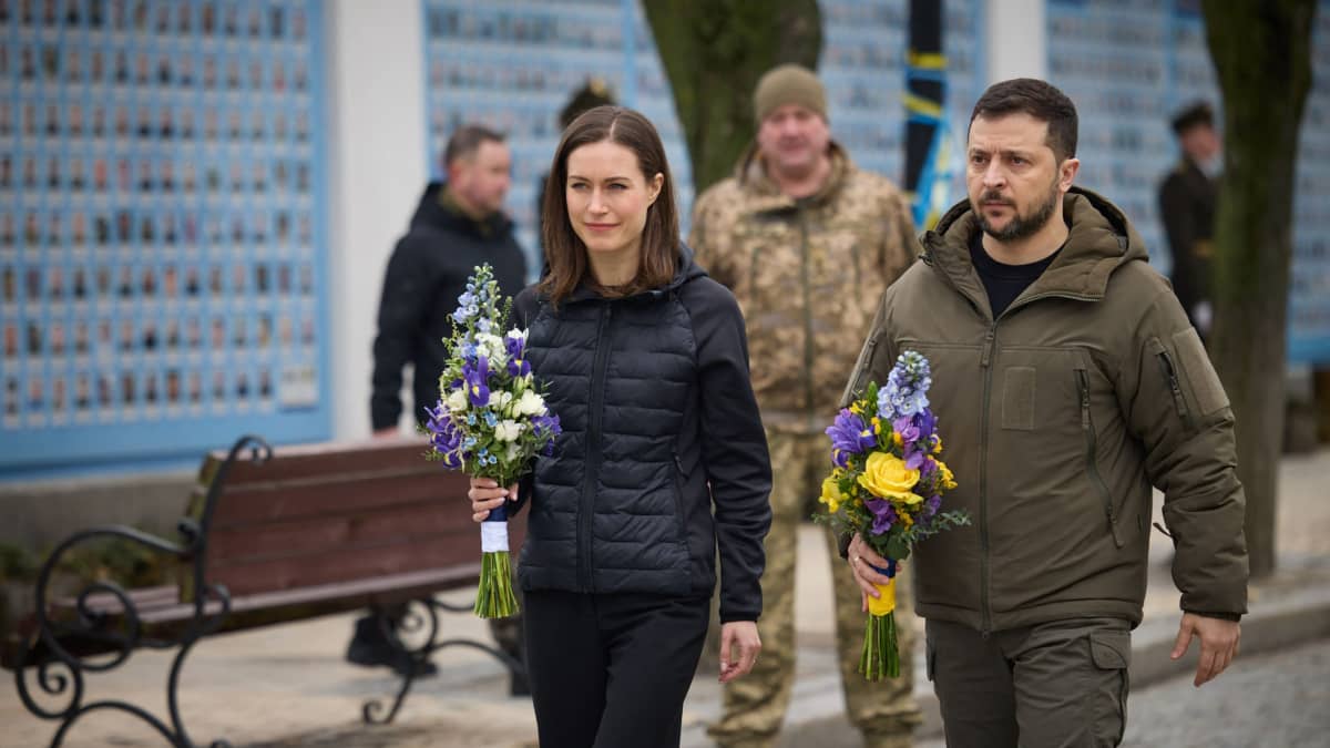 Sanna Marin ja Volodymyr Zelenskyi kävelevät rinnakkain kukka kimput kädessä kadulla. Taustalla sotilas asuinen henkilö ja ohikulkijoita.