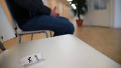 Etualalla rypistynyt vuoronumerolappu valkoisella tuolilla, taustalla henkilö farkut jalassa terveyskeskuksen aulassa.