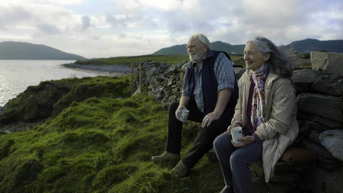 En leende Howard (James Cosmo) och Annie (Bríd Brennan) sitter vid en stenmur och ser ut över havet, bakom dem syns gröna vidder och berg.