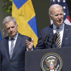 President Biden raises his hands as he speaks at a podium with Niinistö standing just behind him against a backdrop of Swedish, American and Finnish flags.