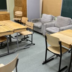 Vanhan luokkahuone, jossa pulpetteja, tuoleja, pari nojatuolia ja opettajan pöytä.