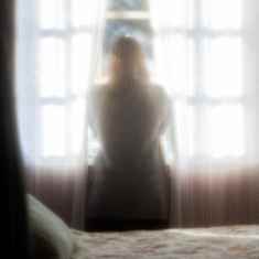 Nainen katsoo verhojen välissä ikkunasta ulos pehmeässä valossa.