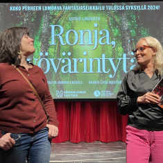 Ohjaaja Liisa Mustonen ja Hämeenlinnan teatterinjohtaja Anna-Elina Lyytikäinen katsovat toisiaan teatterin näyttämön edessä. Taustalla punainen esirippu ja sen edessä näyttö, jossa Ronja ryövärintytär -näytelän mainos.