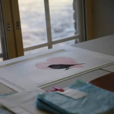 Grafiikkatyö pöydällä, jossa on sydän ja lintu. Maalauksen takana on ikkuna, jossa solisee Tammerkoski.