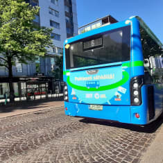 Mukulakivikadulla seisoo takaapäin kuvattu sininen bussi. Bussin perässä on vihreä ja turkoosi mainos, jossa teksti puhtaasti sähköllä. Lehmukset varjostavat katua ja viereistä bussipysäkkiä, vasemmalla näkyy kerrostalo.