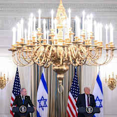 Two men in dark suits stand with serious expressions at podiums in front of US and Israeli flags under a gilt chandelier.