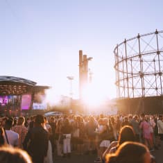 People standing at a festival around sunset with a stage and the frame of an old gasworks in the background.