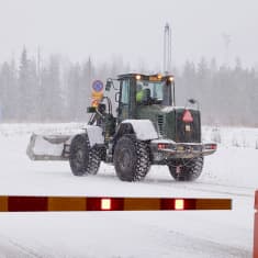 A dark-green tractor carries a concrete blockade in its front-loader in a snowy landscape with a gate saying "border zone" in Finnish.