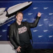 Elon Musk heiluttaa kättään, taustalla pienoismalli avaruusraketista.