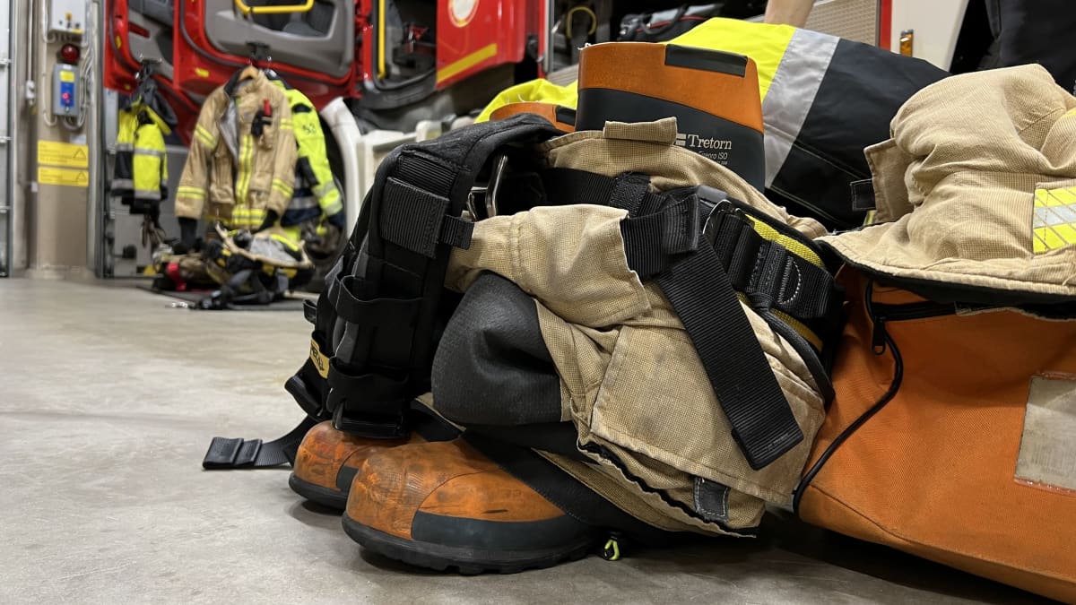 Palomiesten varusteita palolaitoksen autohallin lattialla valmiina hälytystä varten, taustalla paloauto,