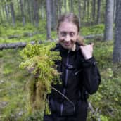 GTK:n erikoistutkija Liisa Maanavilja hymyilee ja pitää kädessään nippua rahkasammalta. Taustalla sammal- ja mustikkapeitteinen kuusikorpi. 