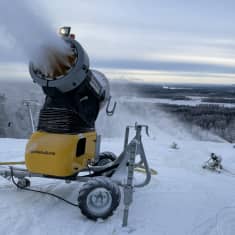 Lumitykki tekee lunta Kasurilan Hiihtokeskuksessa, taustalla rinne ja kolme lumitykkiä. 