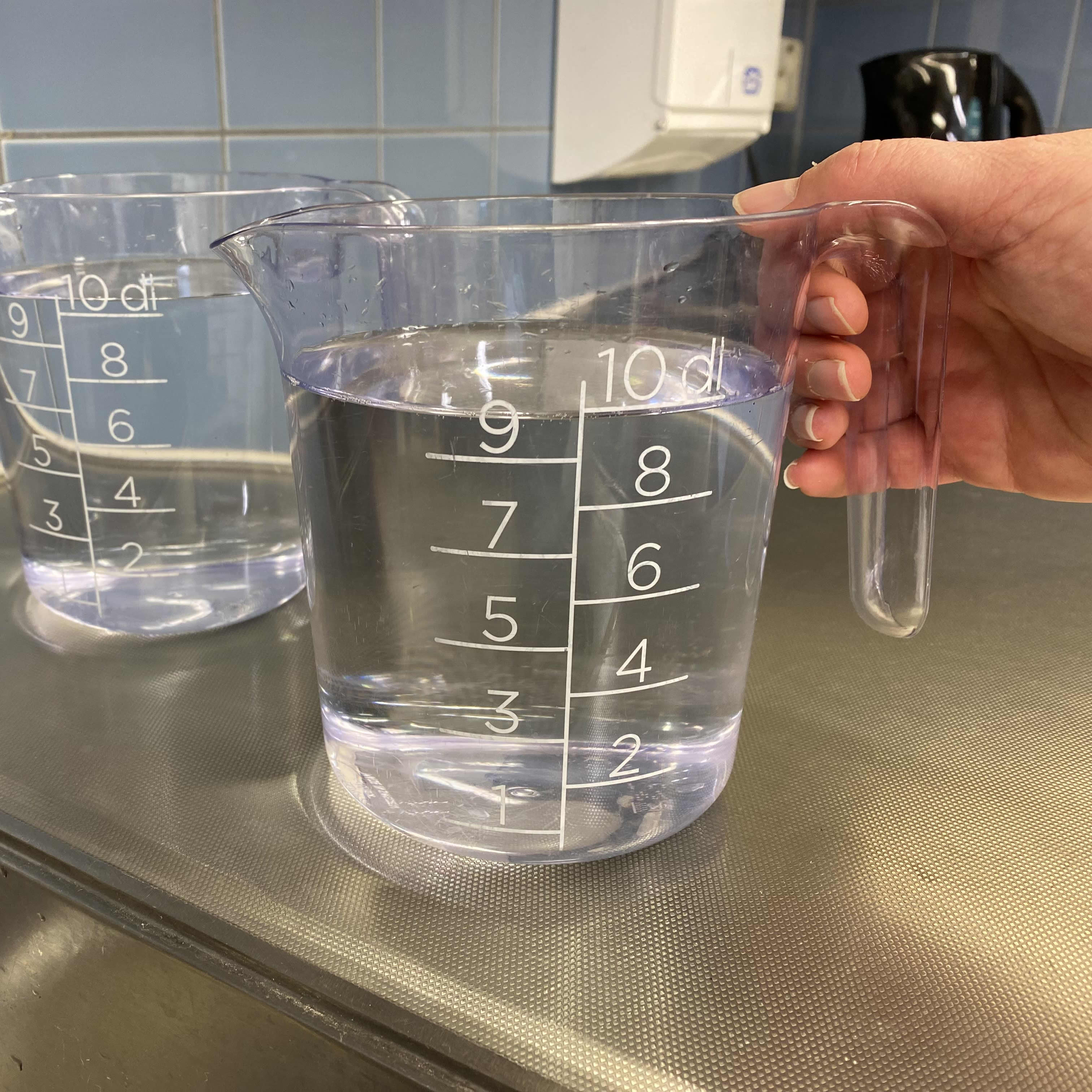Kaksi vedellä täytettyä litran mittakannua. Käsi tarttumassa toiseen niistä.