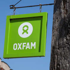 Oxfamin vaaleanvihreä kyltti roikkuu metallikiinnittimistä seinällä, kuvattuna sinistä taivasta vasten.