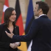 Emmanuel Macron står med ryggen svängd mot kameran medan han pratar med Sanna Marin. Sanna Marin tittar på Macron. I bakgrunden flaggor.