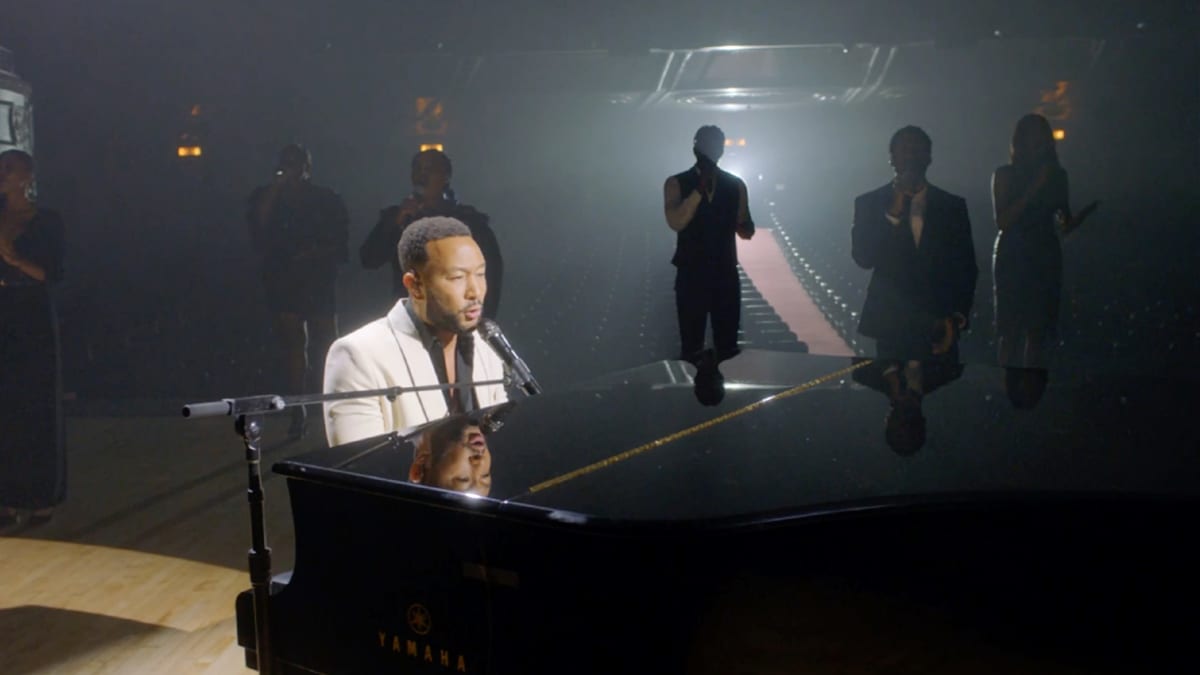 John Legend esiintymässä flyygelin ääressä. Takana taustalaulajia.