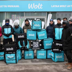 Wolt-lähetit poseeraavat ryhmässä kameralle Wolt-Marketin edessä Kampissa lähettien mielenosoituksen aikana. Edessä myös lähettien ruokareppuja pinottuna.