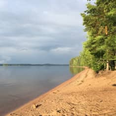 Hiekkarannan rantaviivaa Savitaipaleen Lepänkannon rannassa. Vesi on lähes tyyni, aurinko paistaa mutta taivaalla on tummia pilviä.