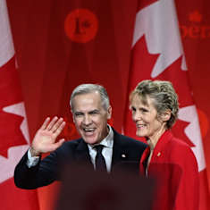 Mark Carney puku päällä vilkuttaa hymyillen, vieressään punaisessa jakussa Diana Carney. Takana Kanadan lippu ja Kanadan liberaalipuolueen logo.