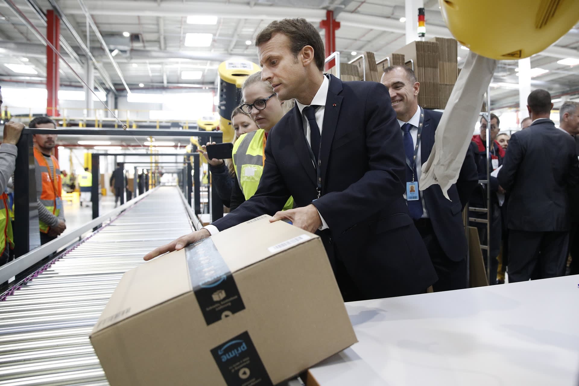Emmanuel Macron nostaa Amazonin paketin liukuhihnalle.
