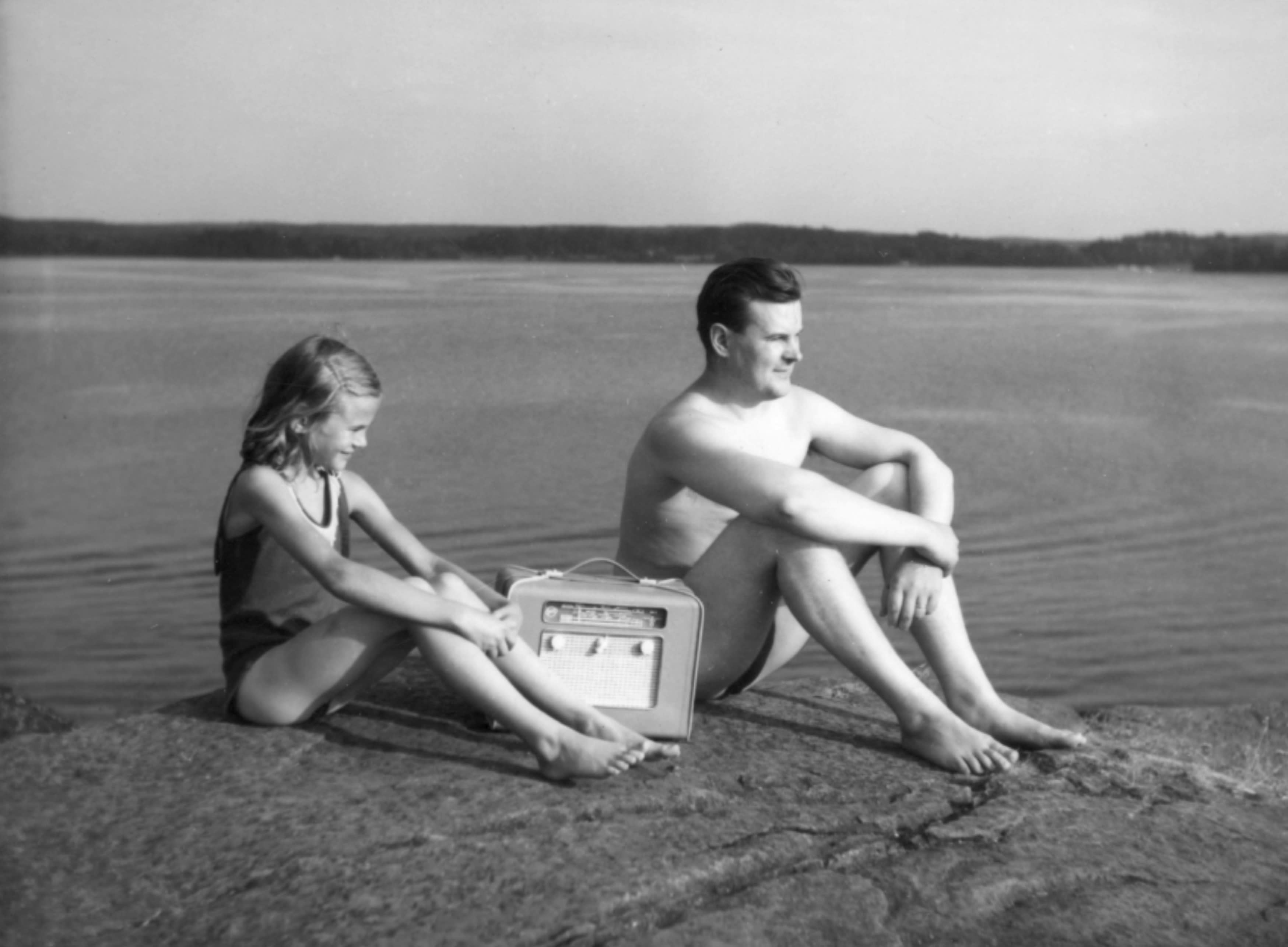 Mies ja lapsi kuuntelevat radiota rantakalliolla, mustavalkoisessa valokuvassa.