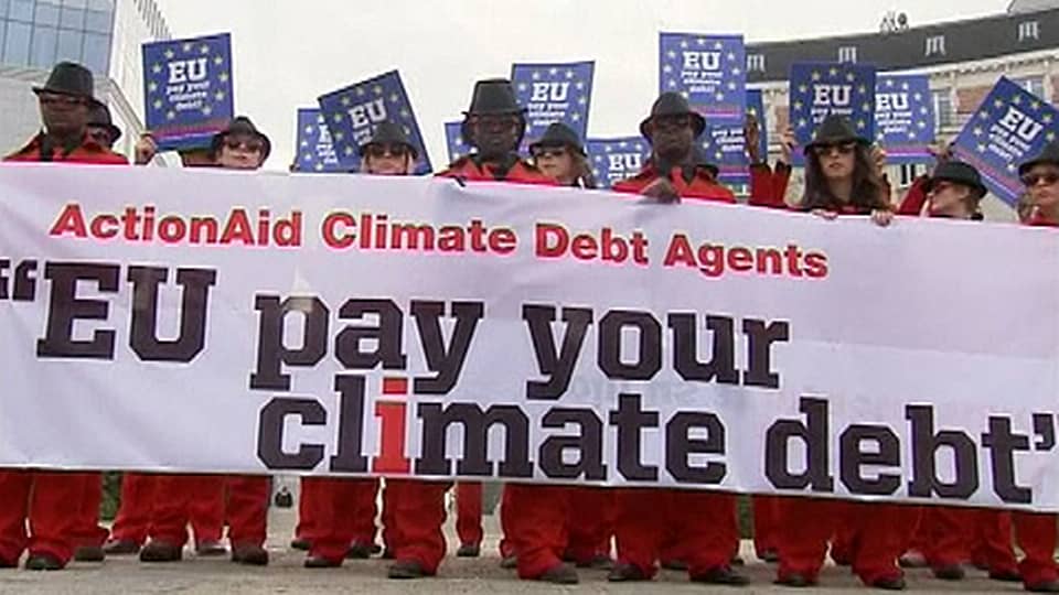 Punaisiin pukuihin ja aurinkolaseihin pukeutuneita mielenosoittajia seisoo rivissä Brysselissä. Pitävät käsissään lakanaa, jossa lukee "EU pay your climate debt".