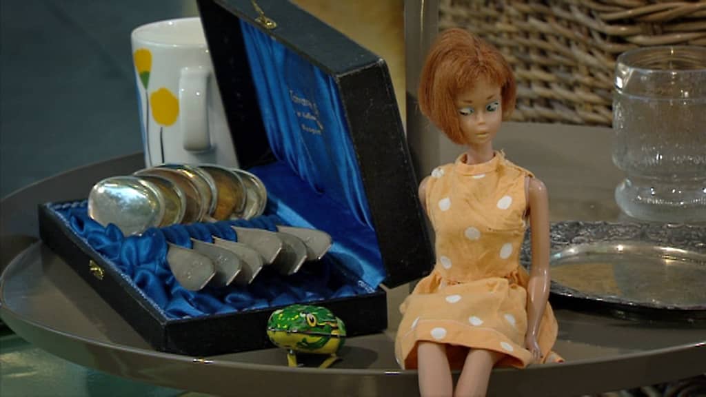 Pop-antiikkia pöydällä. Edessä pikku sammakko ja Barbie-nukke, takana hopealusikoita.