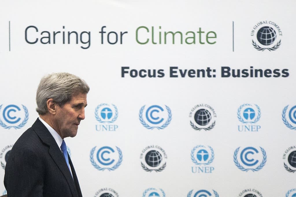 Yhdysvaltain ulkoministeri on kuvan vasemmassa laidassa tummassa puvussa, sininen kravatti kaulassaan. Taustalla on seinätaulu, jossa on tekstiä: "Caring for Climate. Focus Event: Business."