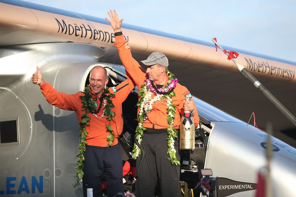 Kaksi iloista lentäjää  kukkaseppeleet kaulassa ja sampanjapullo kädessä viittoilee lentokoneen edessä. 