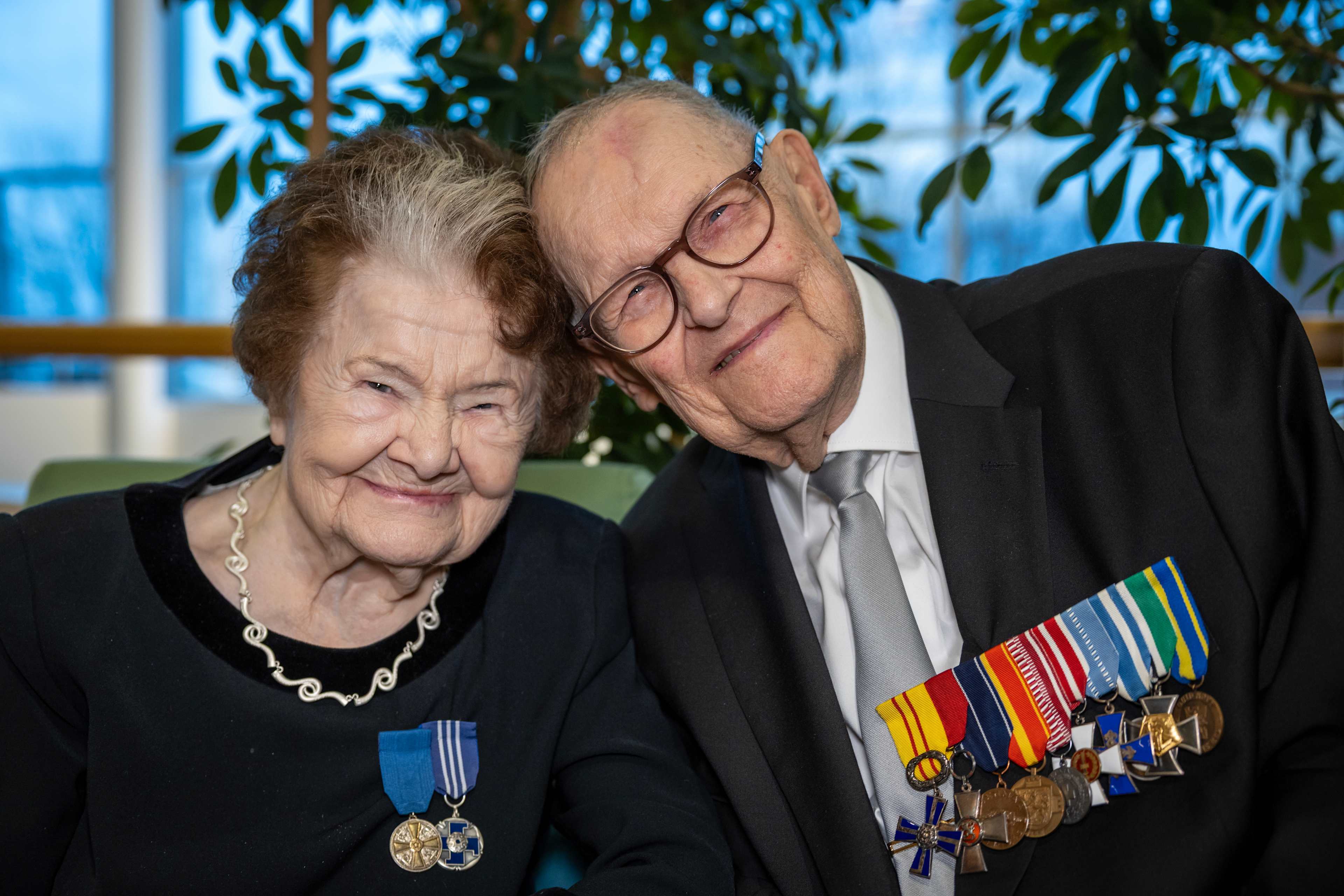 Two veterans, Senni Sopanen and Matti Hara, skating hand-in-hand at a festive ice rink event, their faces lit by the glow of holiday decorations