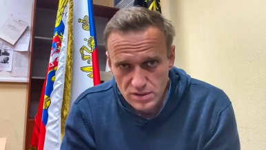 Aleksei Navalnyi katsoo vakavana kameraan. Hänellä on yllään sininen pusero, Taustalla huoneessa näkyy lippuja.ja ilmoitustaulu.