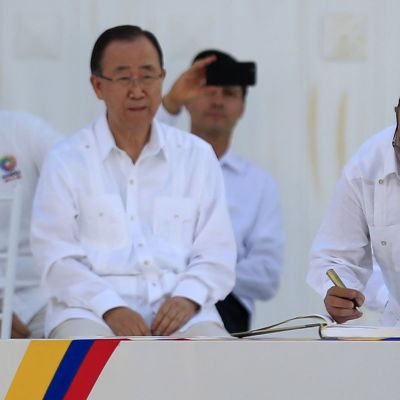 Ban Ki-moon istuu vieressä, kun Rodrigo Londono Echeverri allekirjoittaa sopimusta. Taustalla näkyy muita ihmisiä. Kaikki ovat pukeutuneet valkoiseen.