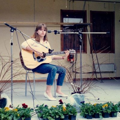En ung Anneli Magnusson sitter på en stol på en scen med gitarr i handen. Mikrofoner står uppställda mot henne och hon ser rakt in i kameran.