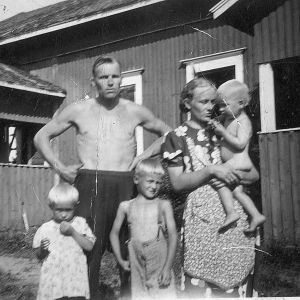 Viisihenkinen perhe puutalon edustalla kesäpäivänä. Perheen isä paidattomana edessään tytär ja poika. Äiti seisoo vieressä pienin lapsi sylissään.
