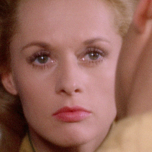 Nainen (näyttelijä Tippi Hedren) lähikuvassa katsoo hieman surumielisen mutta lannistuneen näköisenä suoraan eteensä, ja hänen edessään selin kameraan on mies, josta näkyy hieman päätä ja kaulusta. Kuva Hitchcockin elokuvasta Marnie.
