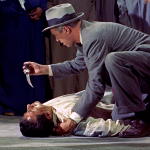 Länsimaiseen pukuun pukeutunut hattupäinen mies (näyttelijä James Stewart) katsoo kädessään olevaa veristä tikaria maassa makaavan murhatun miehen ruumiin ylle kyyristyneenä; taustalla näkyy pohjoisafrikkalaisittain pukeutuneita ihmisiä. Kuva elokuvasta Mies joka tiesi liikaa.