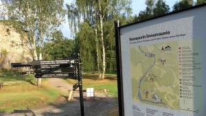 Forststyrelsen skulle resa infotavlor också inne i borgen. Men nu får de vänta på grund av överraskningsfynden. En informationsskylt i färg med en karta och text, en svart stolpe med små svarta skyltar som pekar åt olika håll, slottruinerna i bakgrunden. Sommar, sandväg.