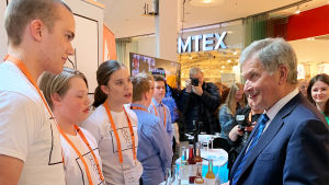 Till vänster synns tre umgdomar som står bakom ett bord med läskedrycker på. Till höger synns president Sauli Niinistö som pratar med dem.
