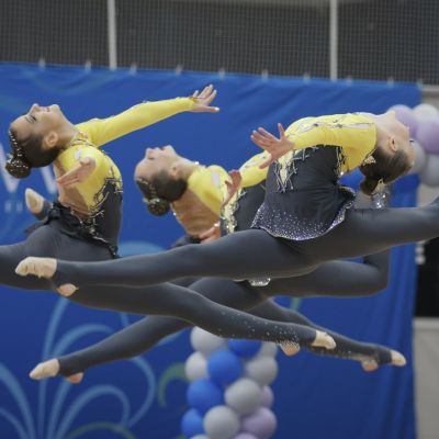 Tre unga kvinnor i gul-grå gymnastikdräkter gör spagat i luften