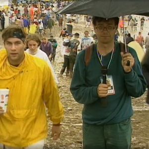Toimittaja seisoo mikki ja sateenvarjo käsissään keskellä sateista festivaalia, vasemmalla keltaiseen sadetakkiin pukeutunut mies kävelee ohi katsoen kameraan. 