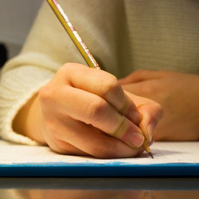 Nuori kirjoitaa paperille lyijykynällä tekstiä. kirjoittaminen kirje käsiala lyijykynä oppiminen lehtiö koulu paperi kello