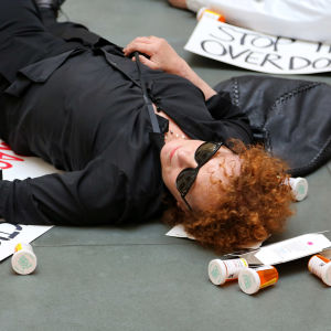 Närbild på Nan Goldin då hon ligger på golvet i ett museum, omgiven av en massa tomma pillerburkar och flygblad.