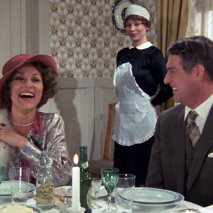 Nauravat nainen ja mies ruokapöydän ääressä, taustalla hymyilevä piika.