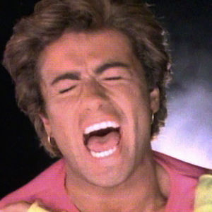 George Michael laulaa silmät kiinni ja hampaat hohtaen Wham!-yhtyeen musiikkividolla. Tumma tausta. Artistilla kirkkaanväriset vaatteet.
