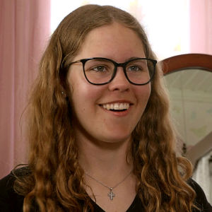 Ung kvinna med långt lockigt hår och glasögon sitter framför ett fönster och ser glad ut.