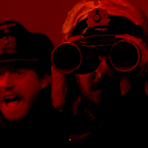 Koppalakkipäinen meriupseeri katsoo kiikareilla kohti kameraa punaisessa tulipalon hehkussa, taustalla kauhistuneen näköisiä miehiä. Kuva elokuvasta Sukellusvene U-96.