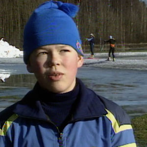 Pipopäinen poika seisoo nurmikentän laidalla, taustalla hiihtäjiä hiihtämässä vielä vähäisellä lumella.