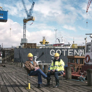 Två män sitter på stolar på en träkaj i en hamn med en kaffetermos.