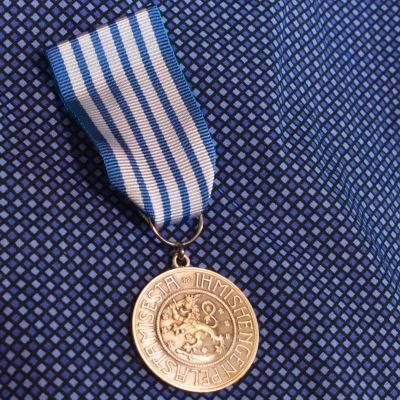 Livräddningsmedaljen i närbild. En rund silvermedlaj med texten "För räddning av människoliv" Medaljen har ett blåvitttt snöre.